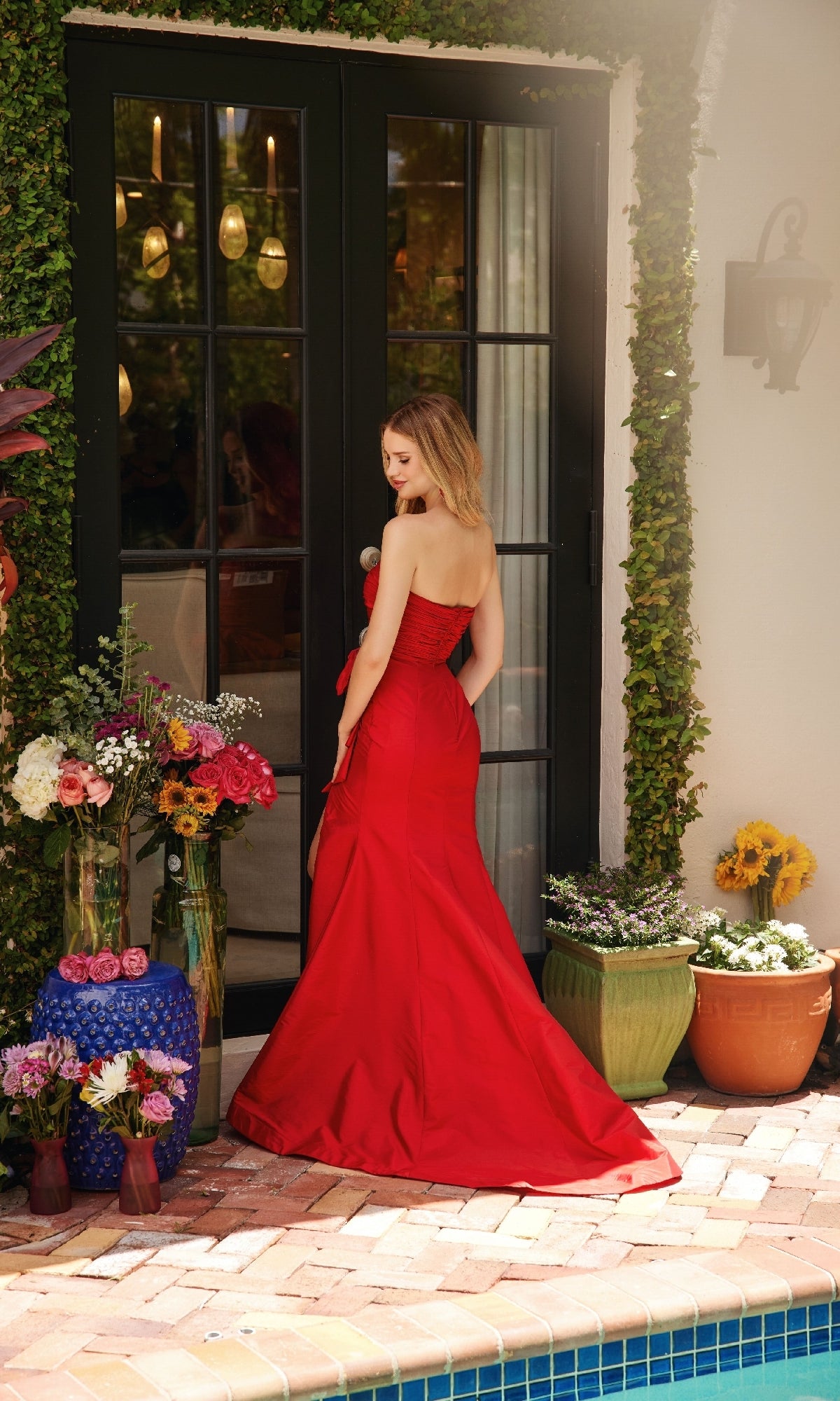 A woman in the Strapless Long Bow Prom Dress: Ava Presley 40072 stands by a glass door, surrounded by potted plants and floral arrangements near a pool.